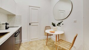 A compact kitchen with a white countertop, a sink, and a stove. There is a dining area with a round table, two wicker chairs, and a potted plant. A large mirror hangs on the wall.