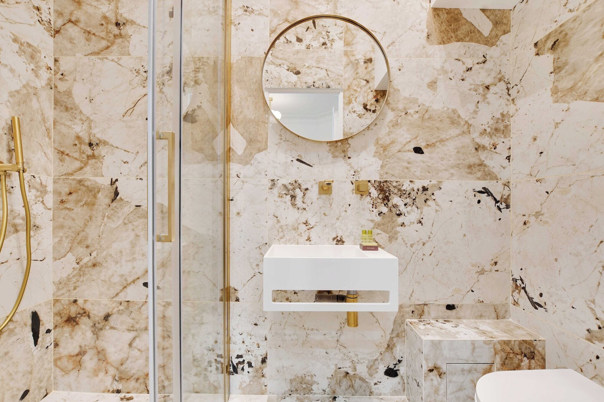 Een badkamer met een marmeren wand, een gouden douche, een witte wastafel en een ronde spiegel.