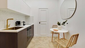 A compact kitchen with a sink, stove, and coffee maker. There is a white door, a round table with chairs, and a potted plant.
