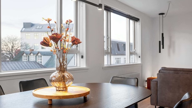 Ein Essbereich mit einem runden Holztisch, einer Vase mit getrockneten Blumen und Blick auf Gebäude durch große Fenster.