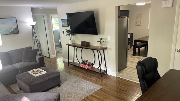 Een woonkamer met een grijze bank, een salontafel, een televisie aan de muur, een eettafel met stoelen en een keuken met een koelkast.