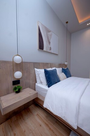 A modern bedroom with a wooden headboard, a bedside table with a plant, and a large bed with white bedding and blue pillows.