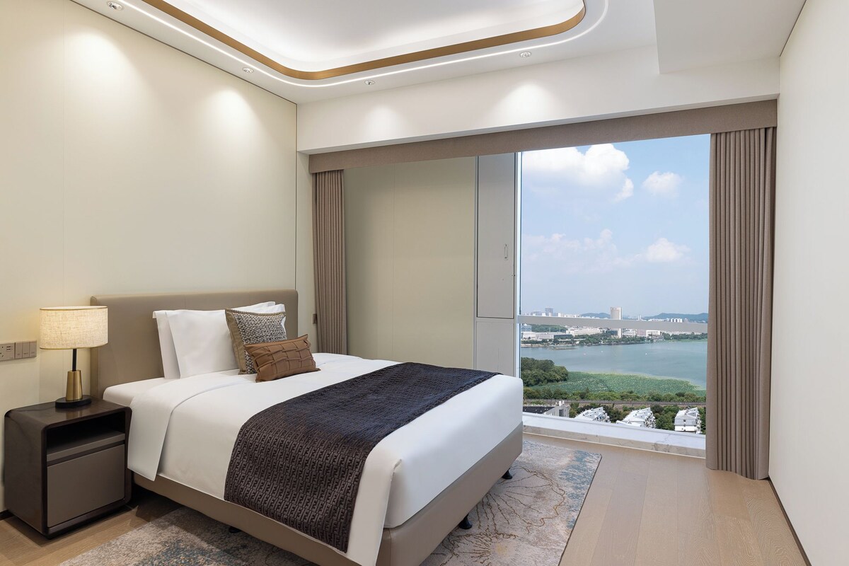 Ein modernes Hotelzimmer mit einem großen Bett, einem Nachttisch, einer Lampe und Blick auf eine Stadtlandschaft und Wasser.