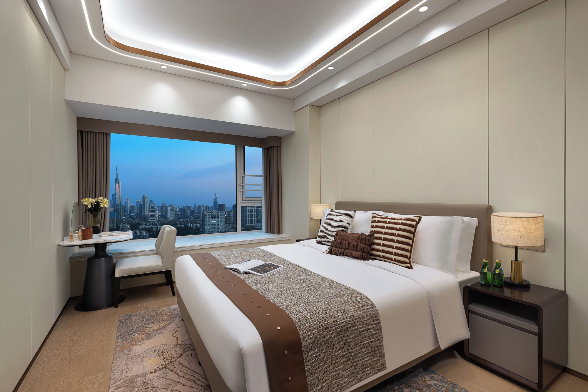 Quarto de hotel com uma cama grande, uma escrivaninha, uma cadeira, uma janela com vista para a cidade e uma mesa de cabeceira com abajur.