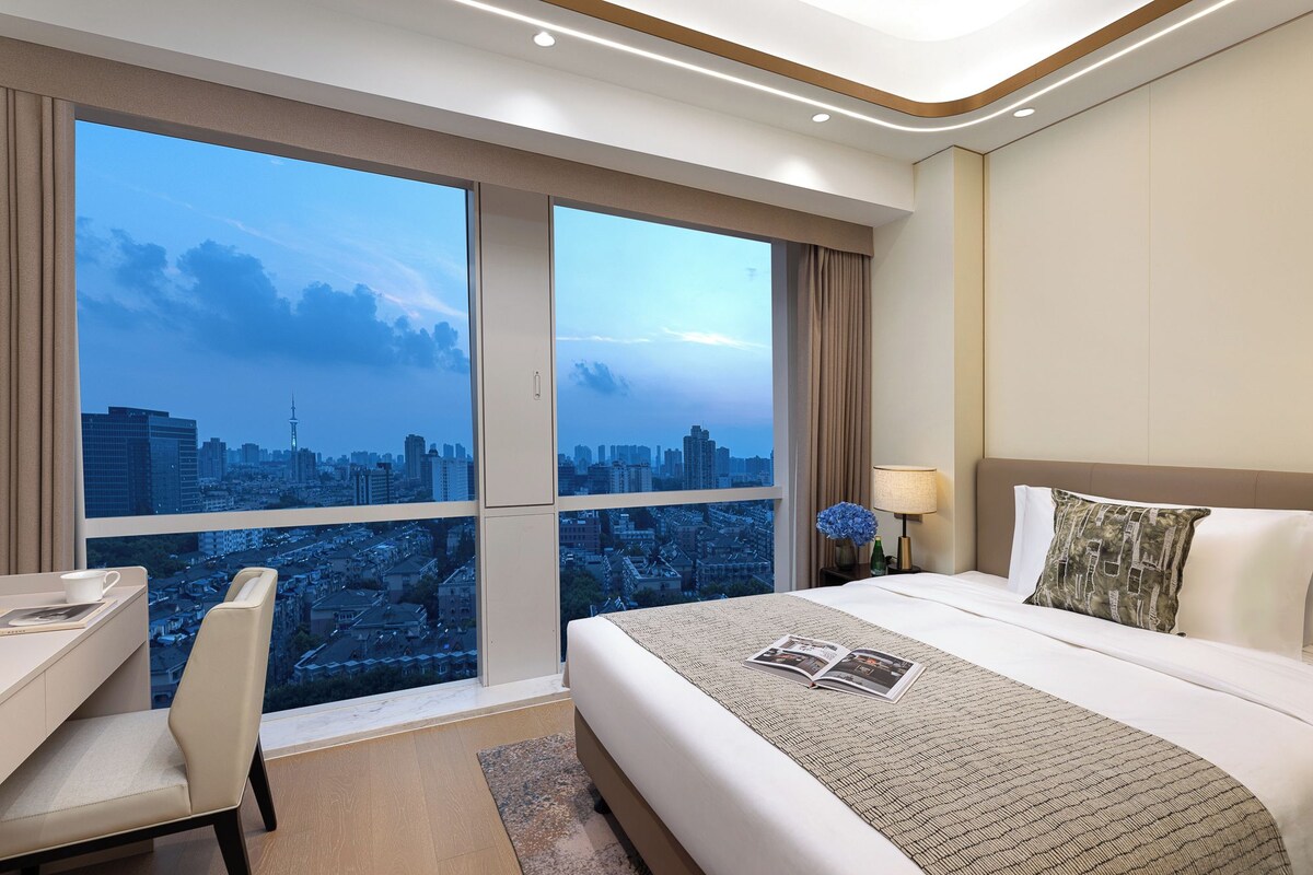 Ein Hotelzimmer mit großem Fenster und Blick auf die Stadt, einem Bett mit gemustertem Überwurf, einem Nachttisch und einem Sessel.
