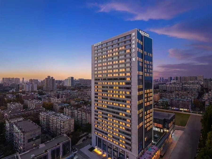 A tall, modern building with the name "ASCOTT" on the facade, surrounded by a dense urban area with various other buildings.