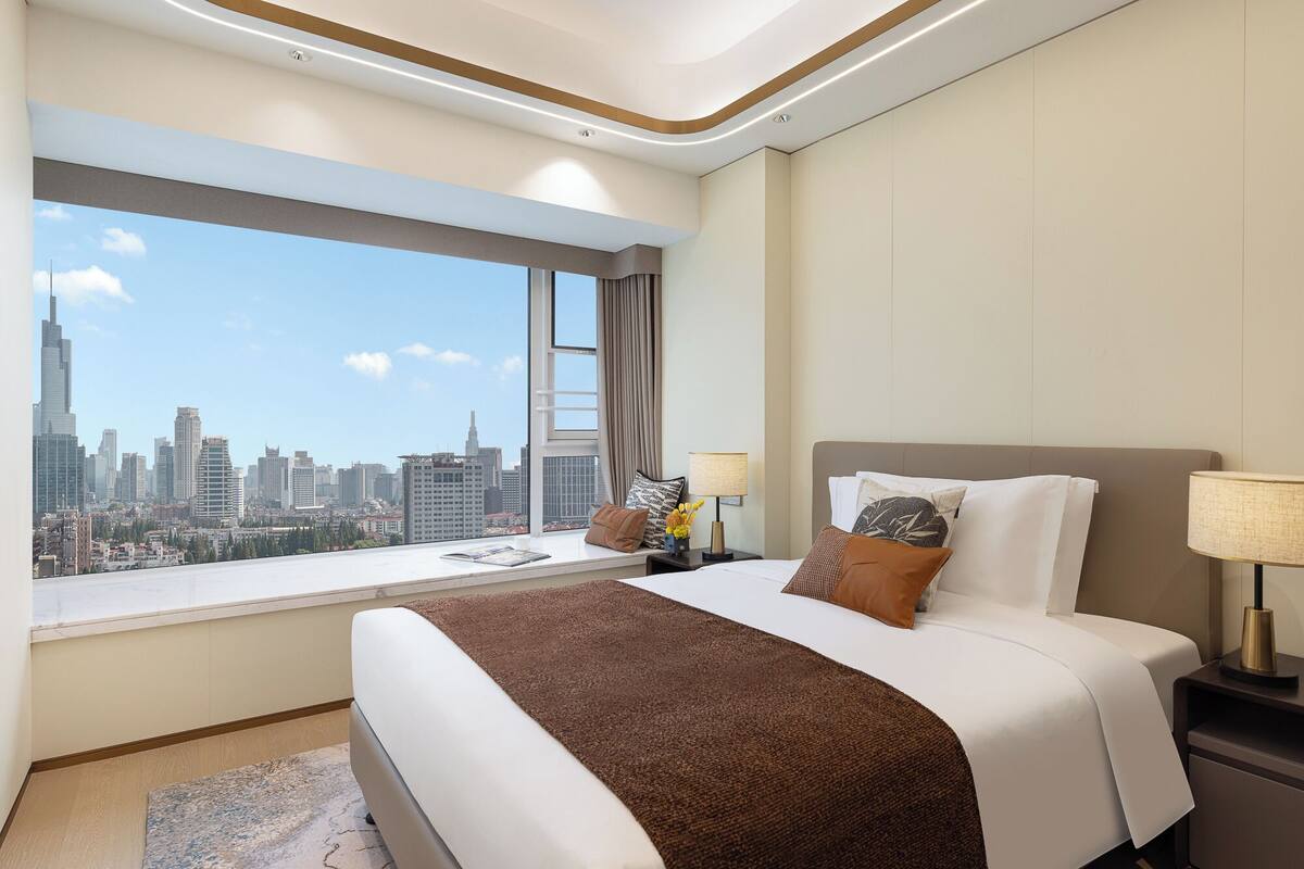 Hotelkamer met een groot raam met uitzicht op de stad, een net opgemaakt bed met een bruine plaid en een nachtkastje met een lamp.