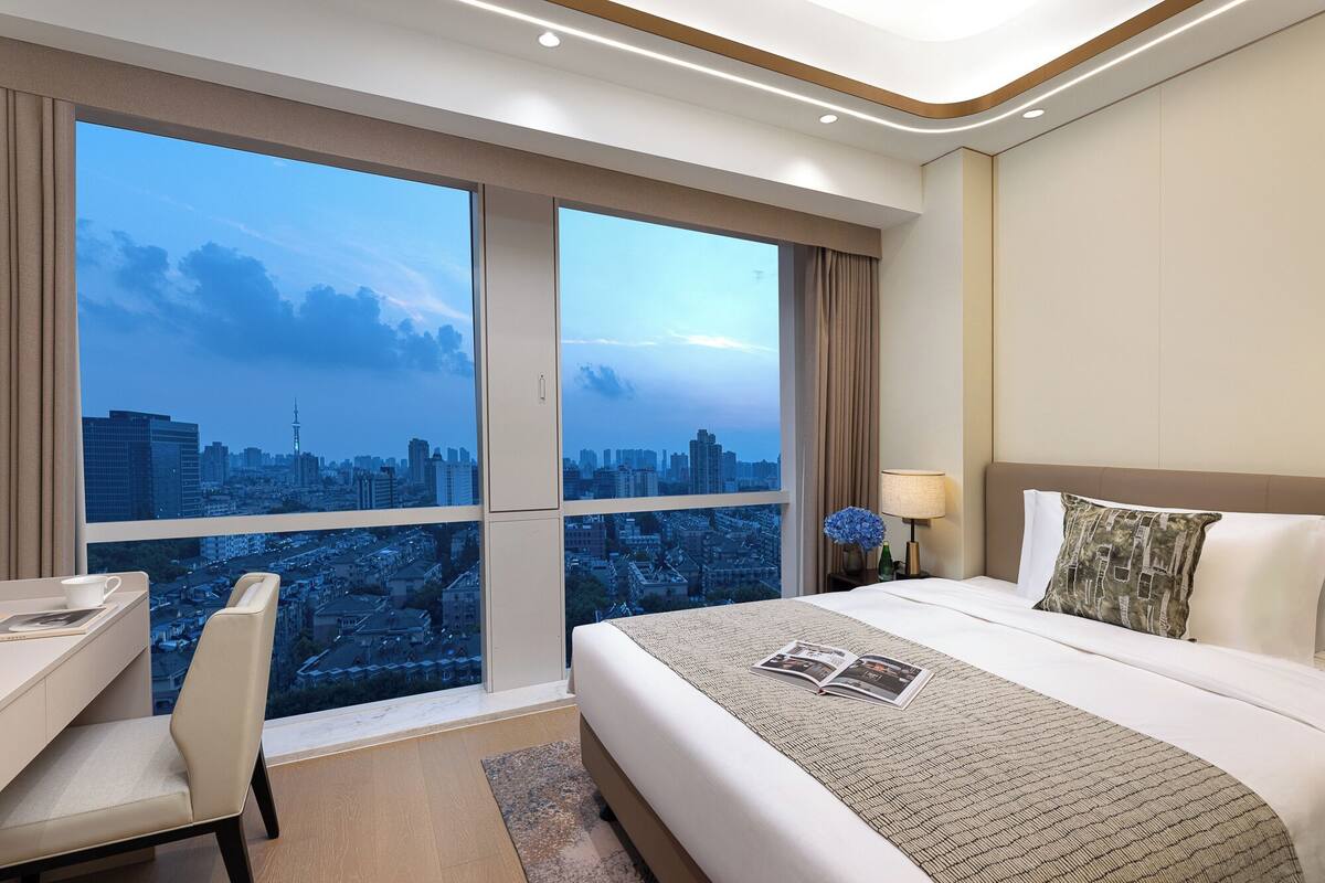 Een hotelkamer met een groot raam en uitzicht over de stad, een bed met een decoratief sprei, een nachtkastje en een stoel.
