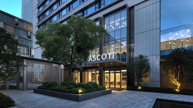 En modern byggnad med en stor glasfasad, en tydlig skylt med texten ”ASCOTT” och ett välskött utomhusområde med planteringar och en gångstig.