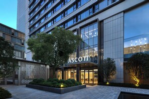 Een modern gebouw met een grote glazen gevel, een opvallend “ASCOTT” bord en een verzorgd buitengebied met plantenbakken en een pad.