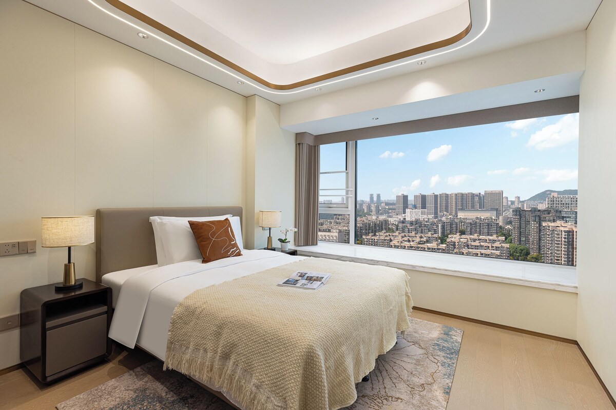 Ein modernes Hotelzimmer mit großem Fenster und Blick auf die Stadt, einem ordentlich bezogenen Bett und einem kleinen Nachttisch mit Lampe.