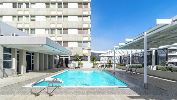 Ein Schwimmbecken mit klarem, blauem Wasser, umgeben von einer steinbelagten Terrasse und einem überdachten Sitzbereich mit Stühlen und Blumentöpfen.