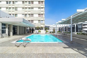A swimming pool with a clear blue water, surrounded by a stone-tiled deck and a covered seating area with chairs and potted plants. - 1 Bedroom Apartment, Seapoint, Cape Town (Cape Town)