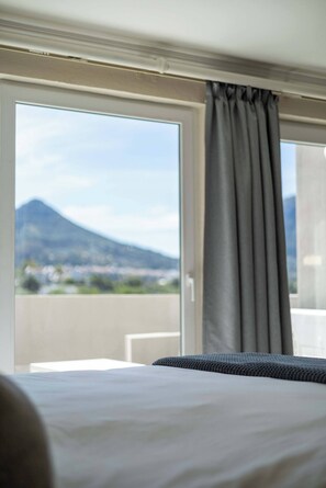 Ett hotellrum med en säng, ett fönster med gardiner och utsikt över ett berg och en stadsbild.