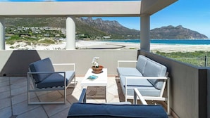 Un balcon offrant une vue sur une plage et des montagnes, avec deux chaises longues, une table ornée d’un vase de fleurs et un livre.