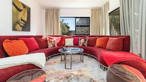 A modern living room with a curved red sofa, a glass coffee table, and a large abstract painting on the wall. - Charming Garden Cottage, Campsbay, Cape Town (Cape Town)