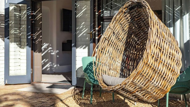 A wicker hanging chair in a covered outdoor area with a green chair and a white wall in the background. - Charming Garden Cottage, Campsbay, Cape Town (Cape Town)