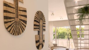 A dining area with a round table and wicker chairs, a white wall with two woven wall art pieces, and a window with a view of greenery.