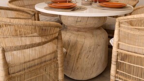 A round dining table with a marble base surrounded by wicker chairs, set with orange plates and wine glasses.