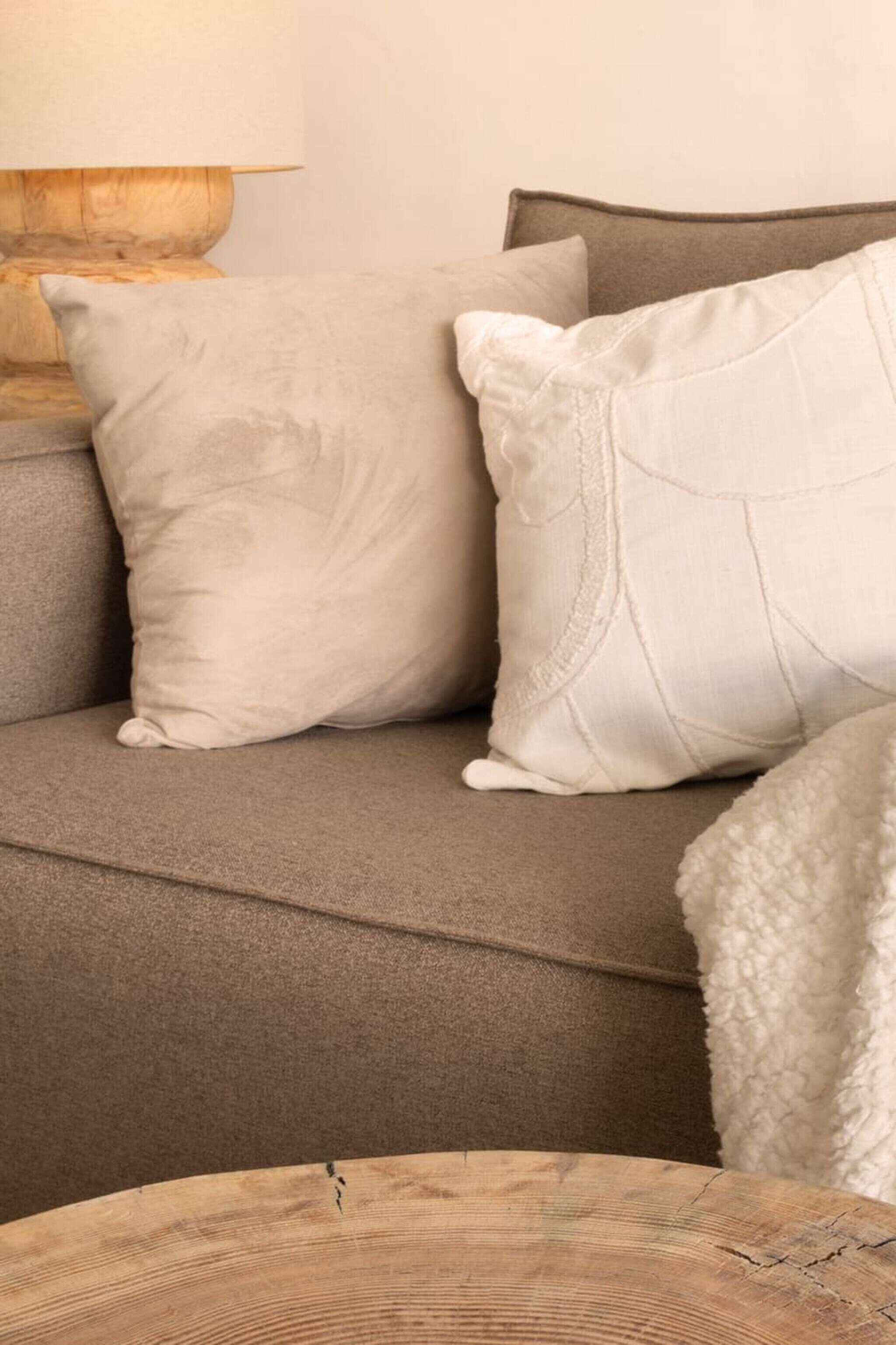 Un canapé beige agrémenté de deux coussins, une table d’appoint en bois et un plaid blanc à texture intéressante.