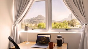 A room with a laptop on a desk, a coffee maker, and a view of a mountain through a window.