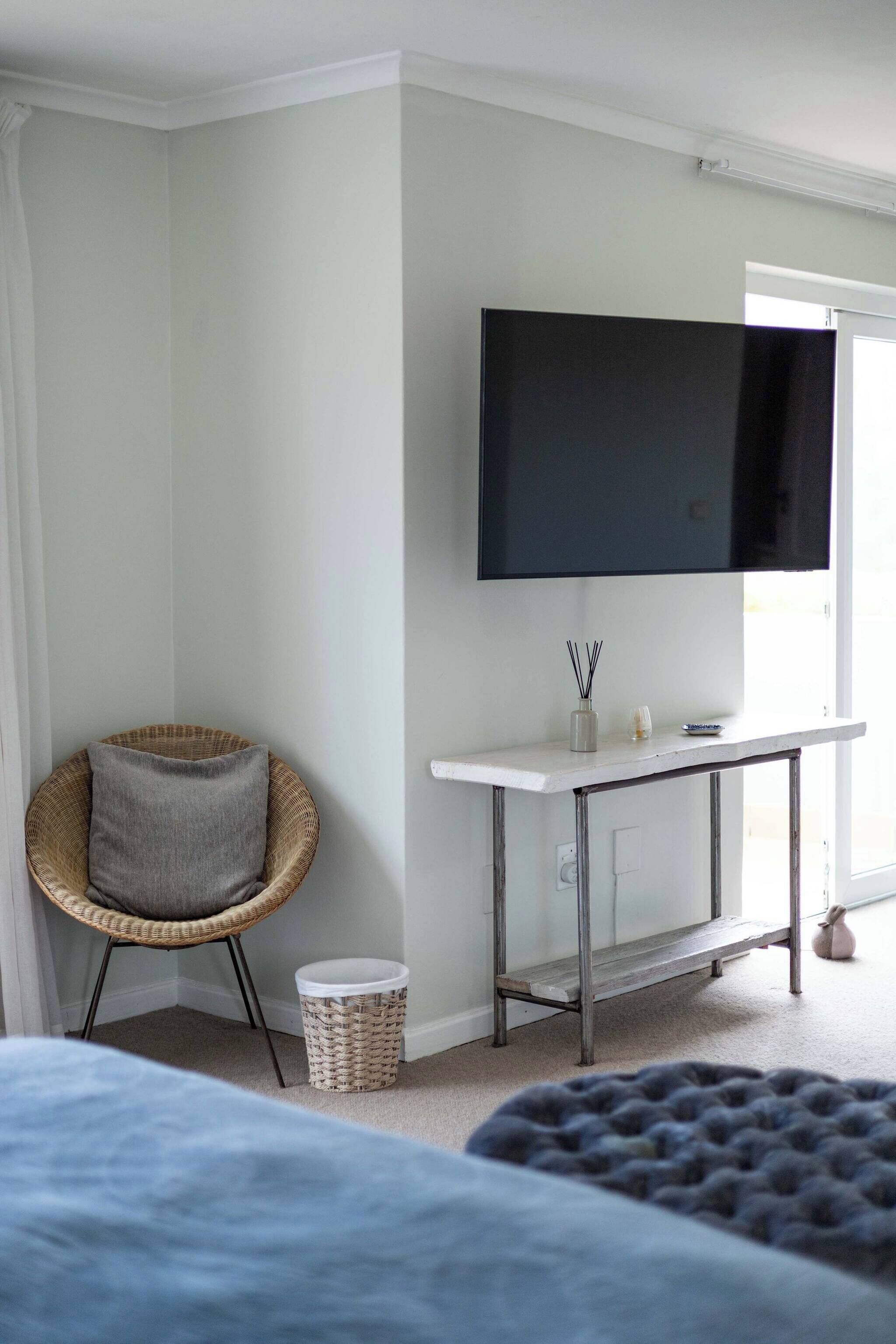 A room with a bed, a wicker chair, a small table with a TV, and a waste bin.