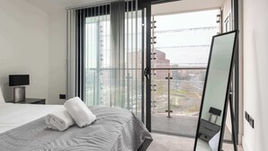 Een moderne hotelkamer met een groot bed, uitzicht op een stadslandschap door middel van kamerhoge ramen, en een spiegel aan de muur.