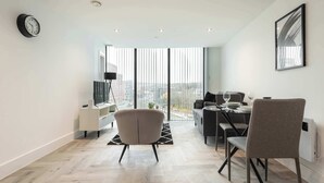 Een moderne woonkamer met een eethoek, grote ramen, een klok en een ingelijst schilderij aan de muur.
