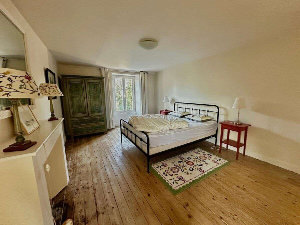 A bedroom with a wooden floor, a bed with white bedding, a small bedside table, a nightstand, a lamp, a dresser, and a window with curtains.