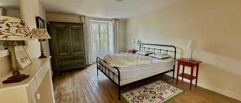 A bedroom with a wooden floor, a bed with white bedding, a small bedside table, a nightstand, a lamp, a dresser, and a window with curtains.
