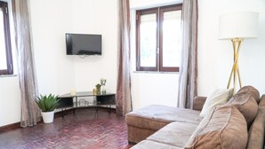 Ein Wohnzimmer mit einer Couch, einem an der Wand montierten Fernseher, einem Esstisch und einer Topfpflanze.