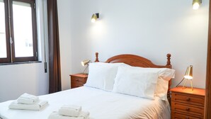 Ein ordentlich bezogenes Bett mit weißen Laken und zwei Kissen, ein Nachttisch aus Holz mit einer Lampe sowie ein Fenster mit Vorhängen.