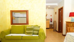 A green sofa with a white throw blanket and pillows, a wooden-framed window, a wooden table with a red lamp, and a tiled floor. - Sacada Apartment - Great Balcony (Guia)