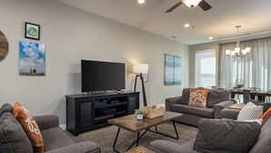 A modern living room with a flat-screen TV, dark wood entertainment unit, grey sofas, and a dining area with a chandelier.