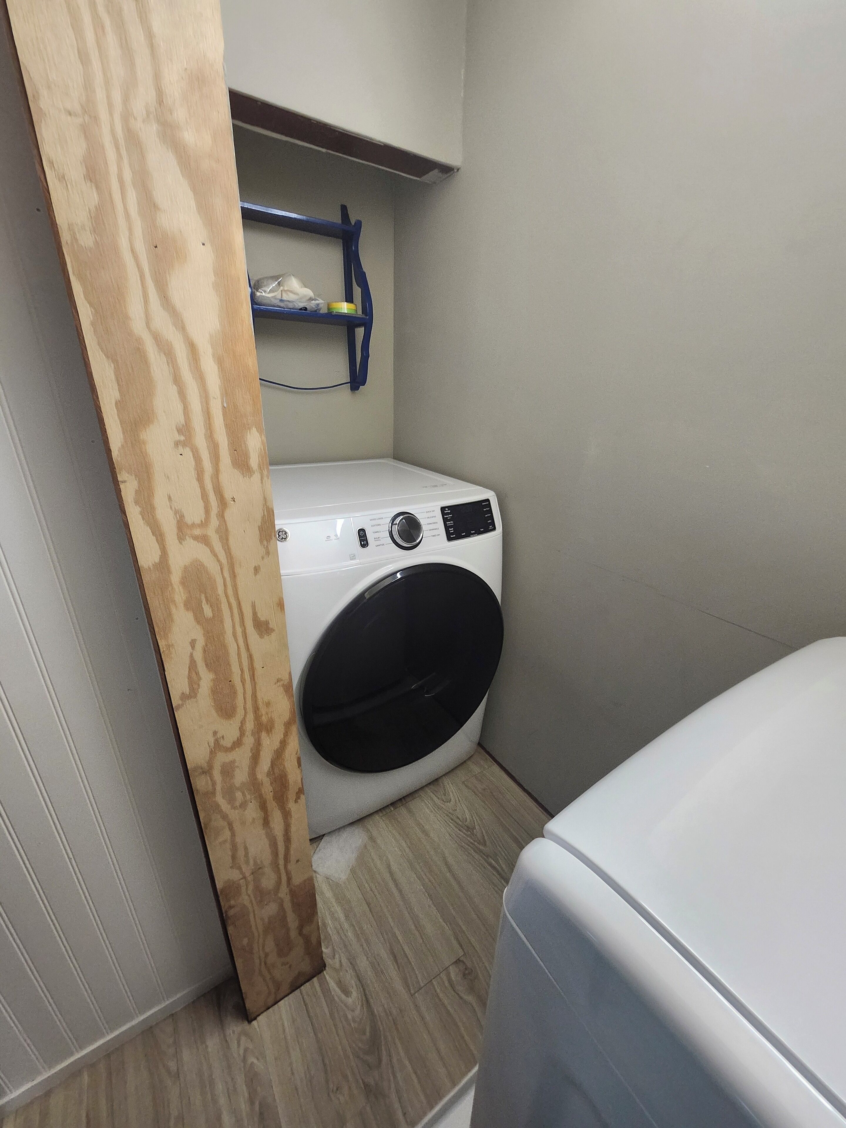 A compact laundry room with a front-loading washing machine, a wooden shelf above, and a small storage area.