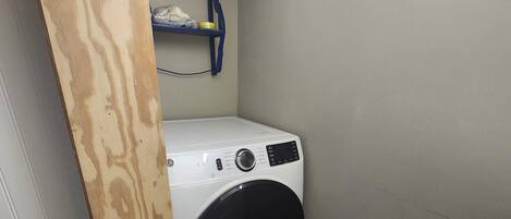 A compact laundry room with a front-loading washing machine, a wooden shelf above, and a small storage area.
