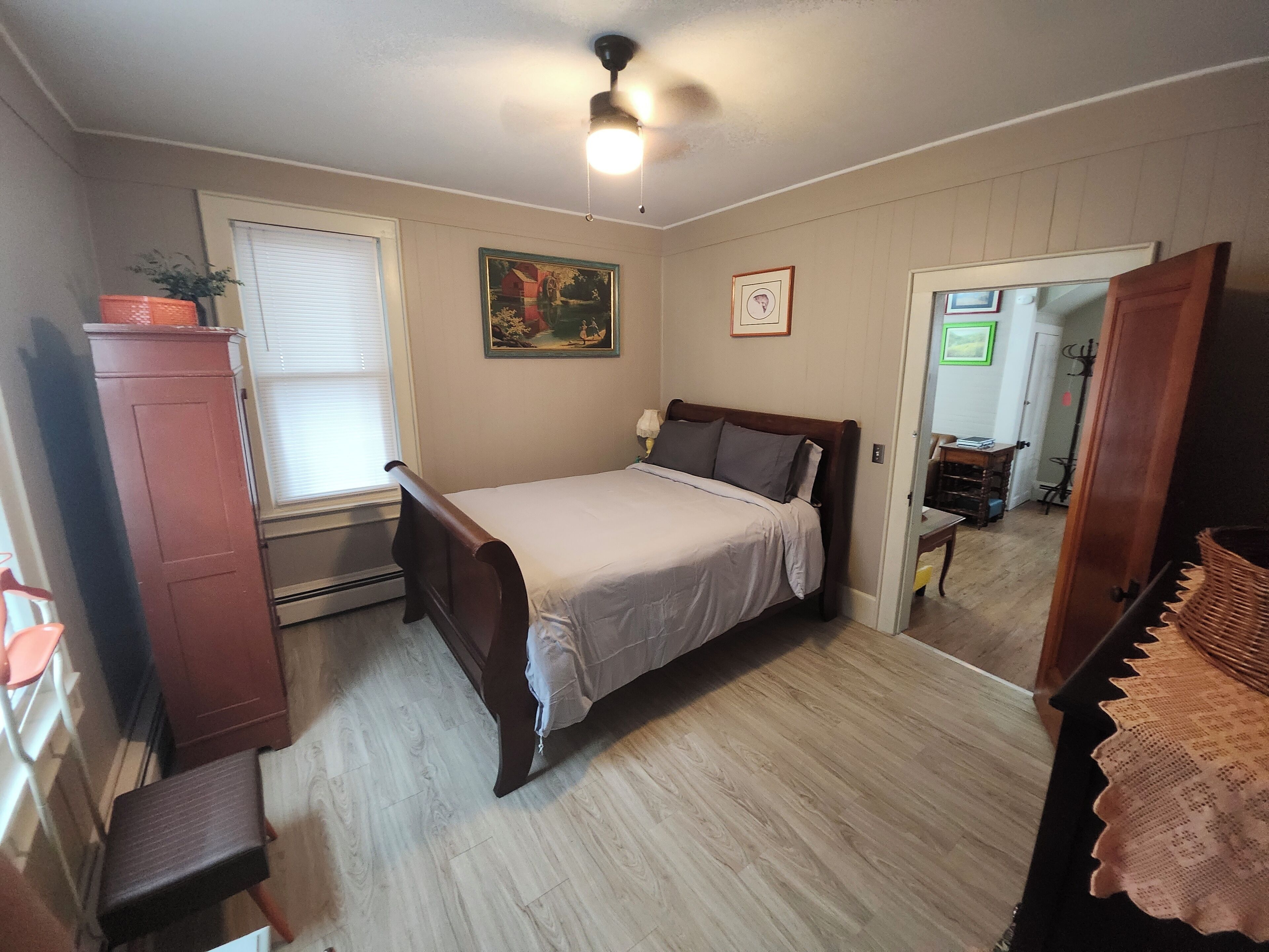 A bedroom with a bed, a dresser, a chair, a window with blinds, a ceiling fan, and a door leading to another room.