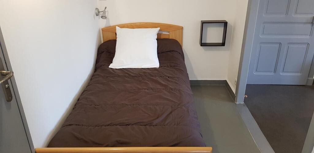 A single bed with a wooden headboard, a white pillow, and a brown bedspread in a room with a gray floor and a white door.