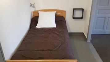 A single bed with a wooden headboard, a white pillow, and a brown bedspread in a room with a gray floor and a white door.