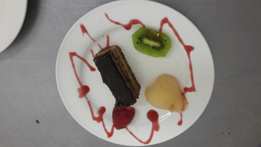 A dessert plate with a slice of cake, kiwi, pear, and strawberry, drizzled with red sauce.