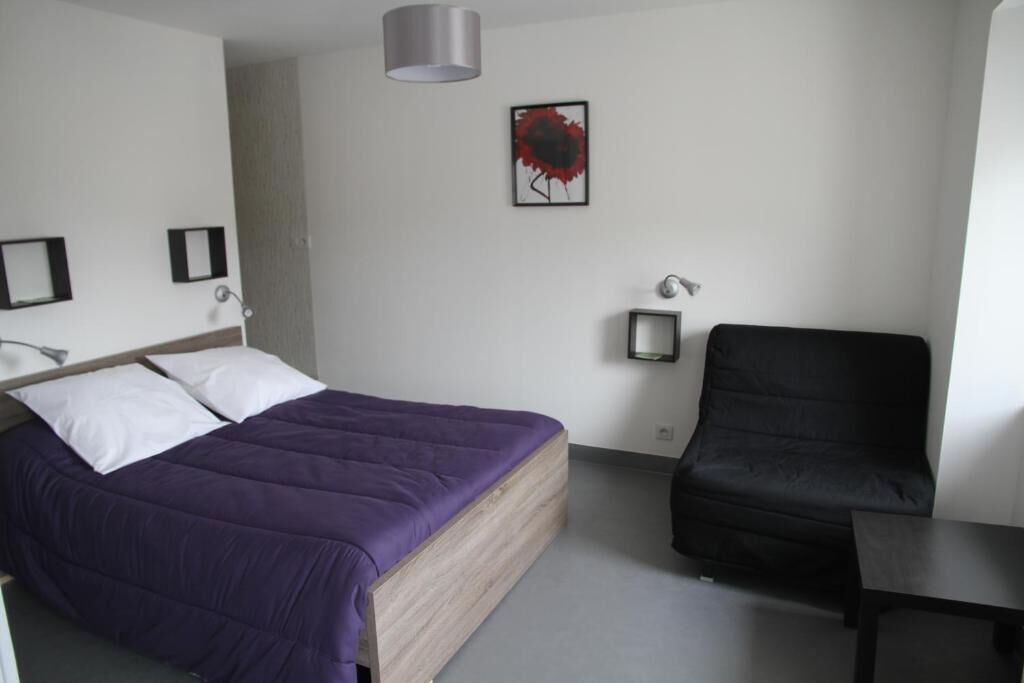 A bedroom with a bed, a black chair, a small table, and a wall-mounted lamp.