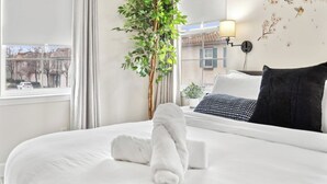 Habitación de hotel con cama, planta en maceta, ventana con cortinas, lámpara de pared y vistas a edificios.