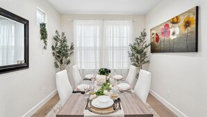 Un comedor con una mesa de madera dispuesta para seis personas, sillas blancas y un cuadro grande en la pared.