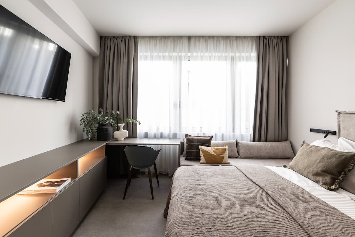 Moderni hotellihuone, jossa on seinälle asennettu taulutelevisio, työpöytä tuolilla, useilla tyynyillä varustettu sänky ja suuri ikkuna, jossa on verhot.