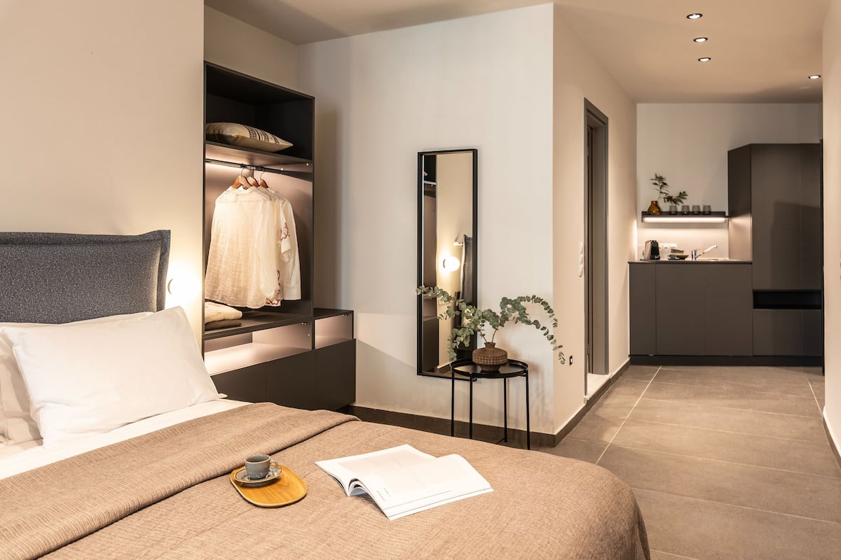 Moderni hotellihuone, jossa on siististi pedattu sänky, yöpöytä, jossa on kirja ja kuppi, peili, pieni sivupöytä, jossa on kasvi, ja taustalla näkyy pieni keittiönurkkaus.