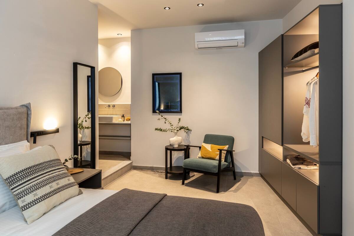 Moderni hotellihuone, jossa on sänky, oleskelualue tuolilla, pieni pöytä, peili ja vaatekaappi.