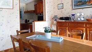 Un coin repas avec une table en bois, des chaises et un vase blanc orné de fleurs. On aperçoit une cuisine en arrière-plan, avec des meubles en bois et un mur recouvert de carreaux.