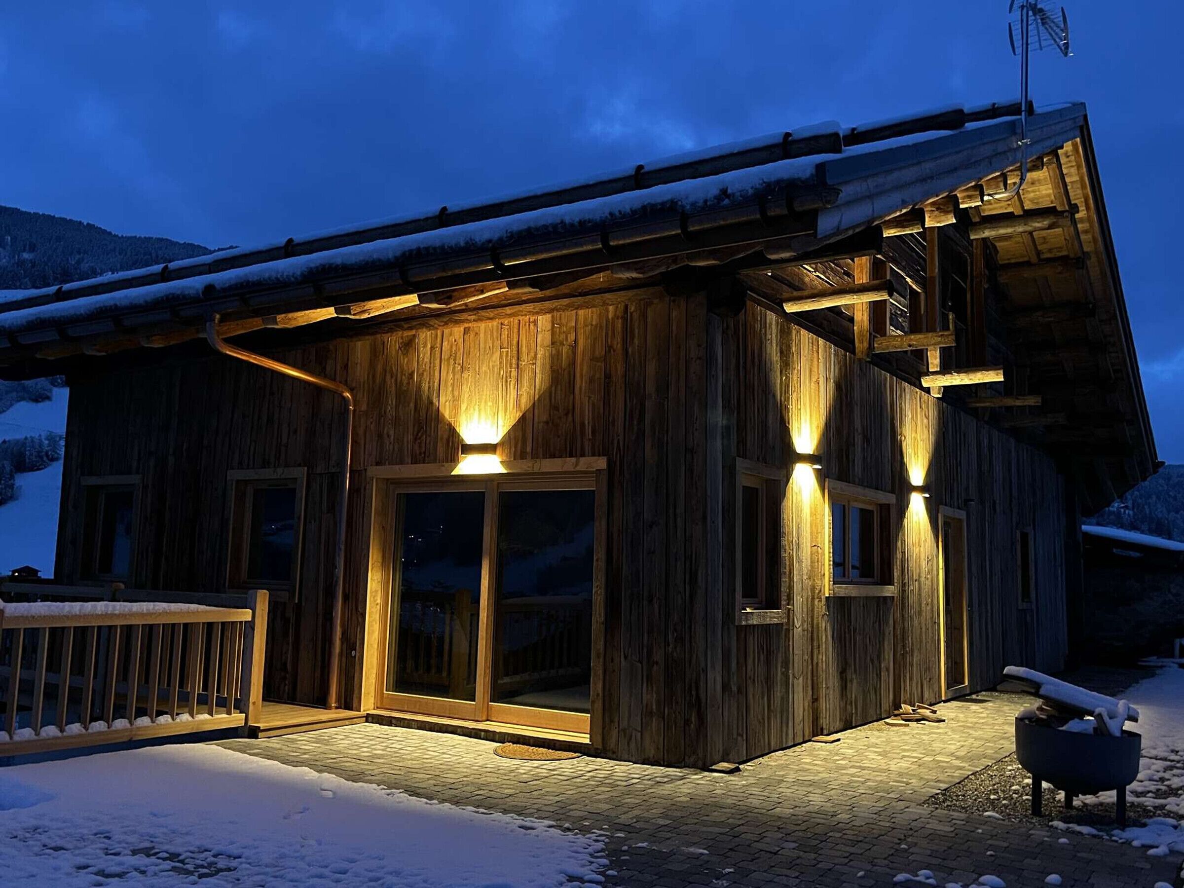 A wooden cabin with a snow-covered roof, large windows, and a wooden deck.