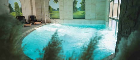 An indoor swimming pool with three lounge chairs, arched windows, and a view of a green landscape.