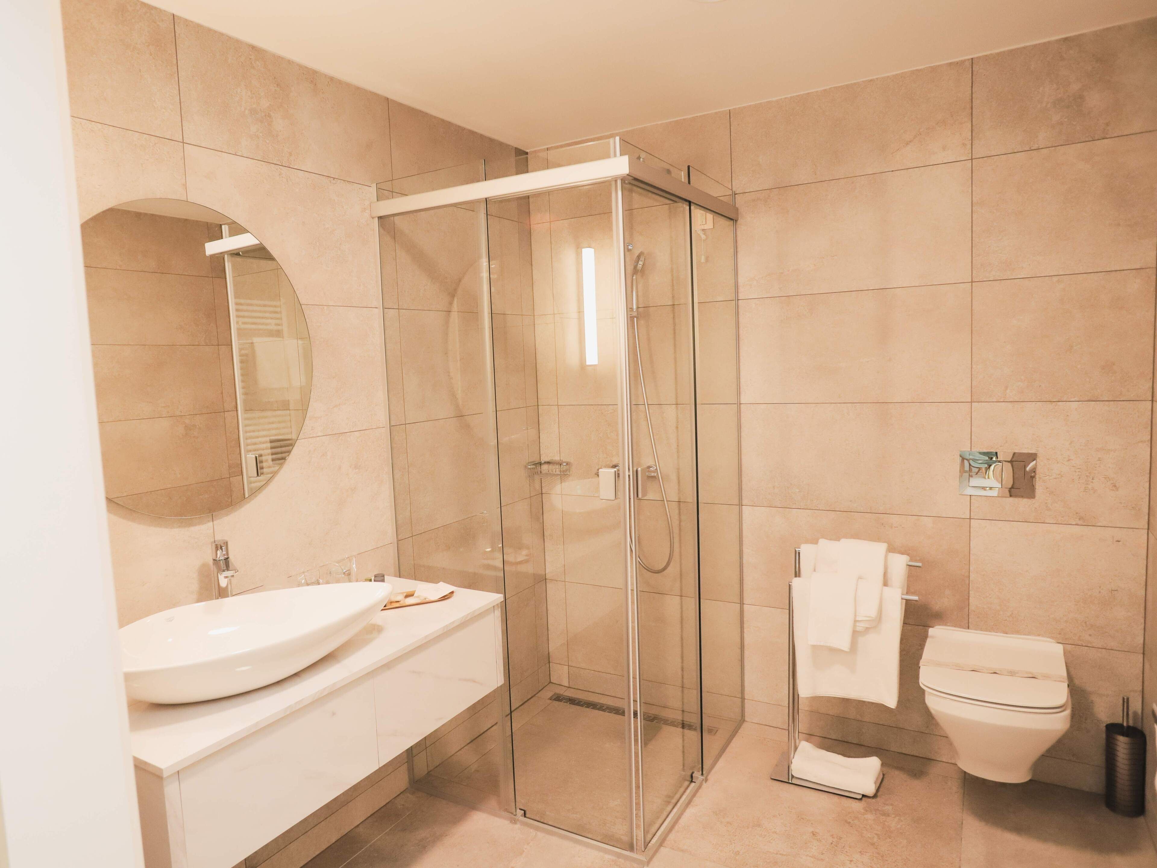 A modern bathroom with a glass-enclosed shower, a large round mirror, a white sink, and a toilet.